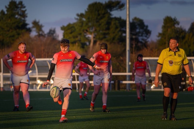 A rugby team in red jerseys and white shorts warming up on a field with a coach or referee in a yellow shirt walking alongside them during sunset.