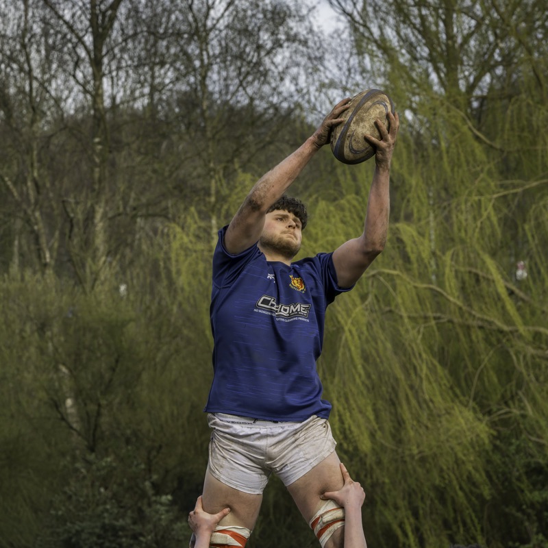 A rugby player in blue jersey lifting a teammate for a line-out in an outdoor field surrounded by trees.