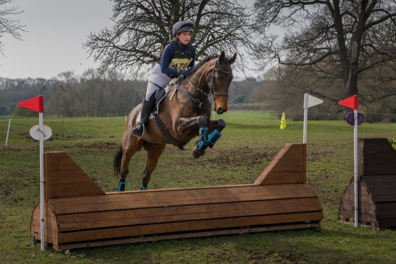 A female equestrian rider dressed in white riding pants, a navy jacket, and a helmet, guides a horse over a wooden obstacle during a cross-country event in an open field with trees in the background.