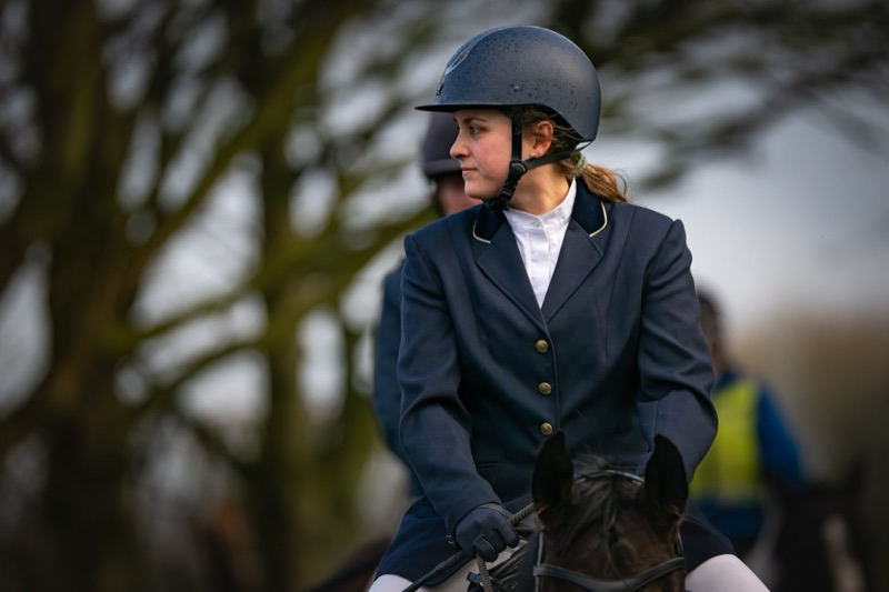 A young woman wearing a riding helmet and navy blazer, sitting outdoors with blurred trees in the background.