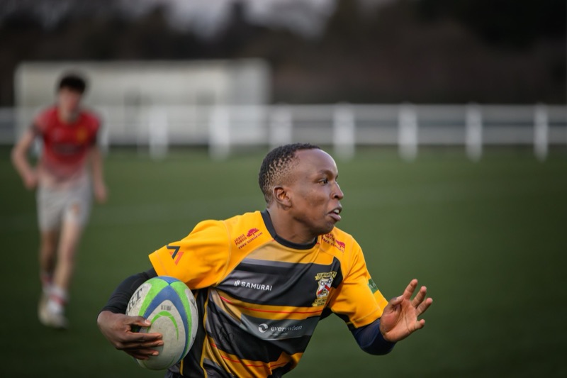 A rugby player in a yellow and black uniform holding a rugby ball in his left hand while running on the field. Another player in a red jersey is blurred in the background near a white fence.