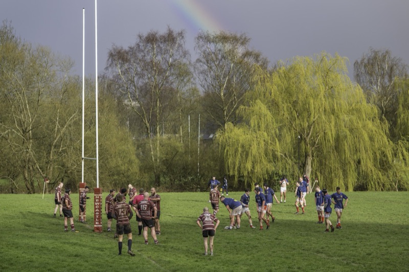 Rugby players in maroon and blue uniforms on a grassy field, with some players standing and others crouching, under a cloudy sky with a rainbow over trees in the background.