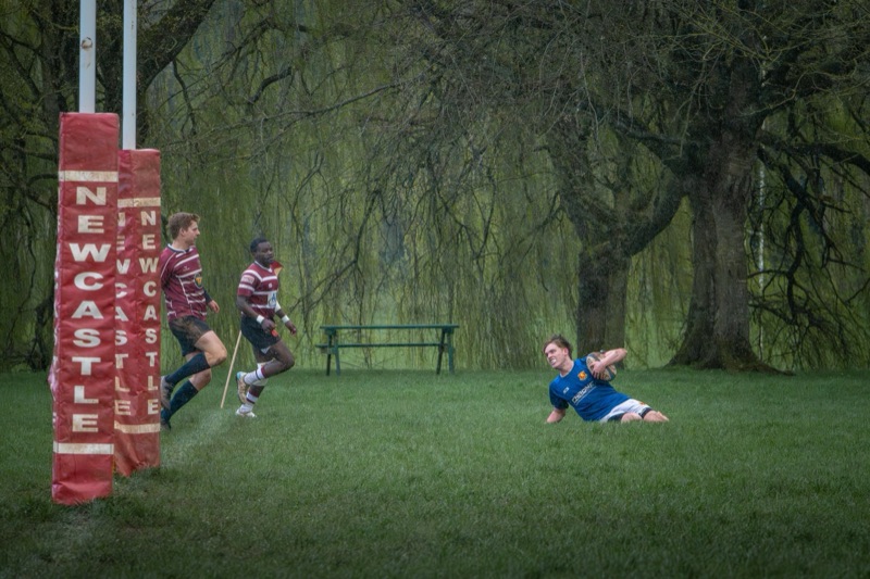 Rugby players in action during a game in the rain, with a player on the ground holding a rugby ball and others running nearby, with a goal post wrapped in red padding and trees in the background.