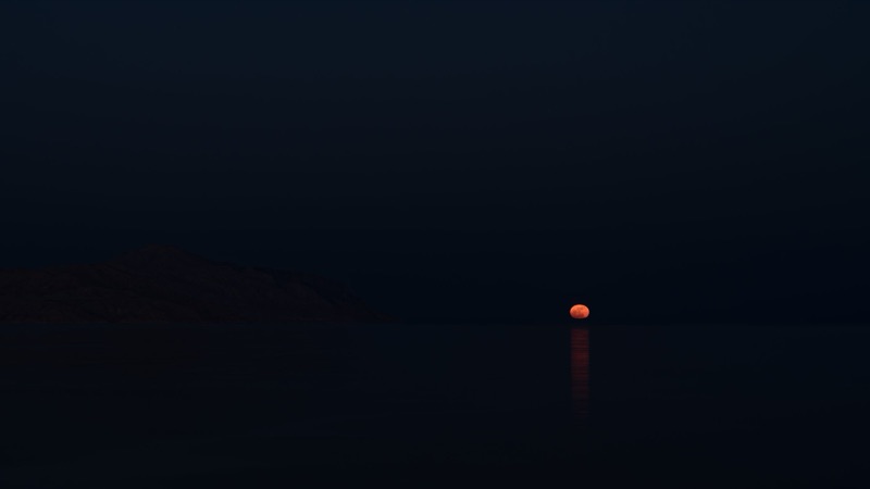 A large reddish full moon rising over a dark horizon with mountain silhouettes and a body of water reflecting the moonlight.