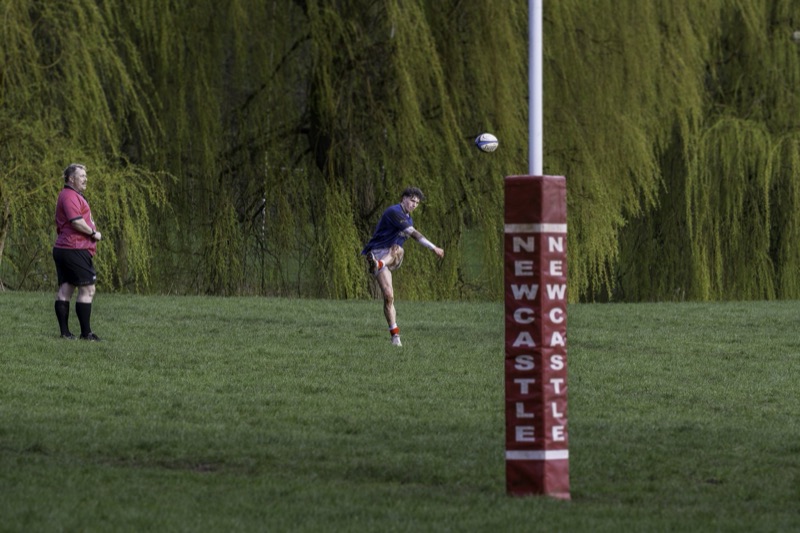 A rugby player kicks a rugby ball towards the goalpost during a game, with another player standing nearby on the field and a lush green background with hanging moss trees.