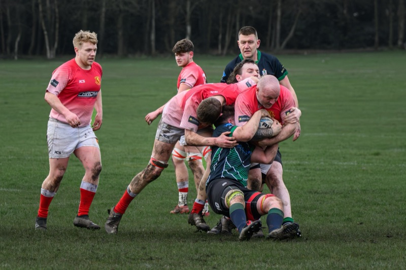 Rugby players in red jerseys tackling their opponent on a grassy field, with some players standing and observing in the background.