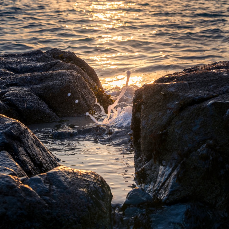 Sunset over a rocky shoreline with water splashing between rocks.