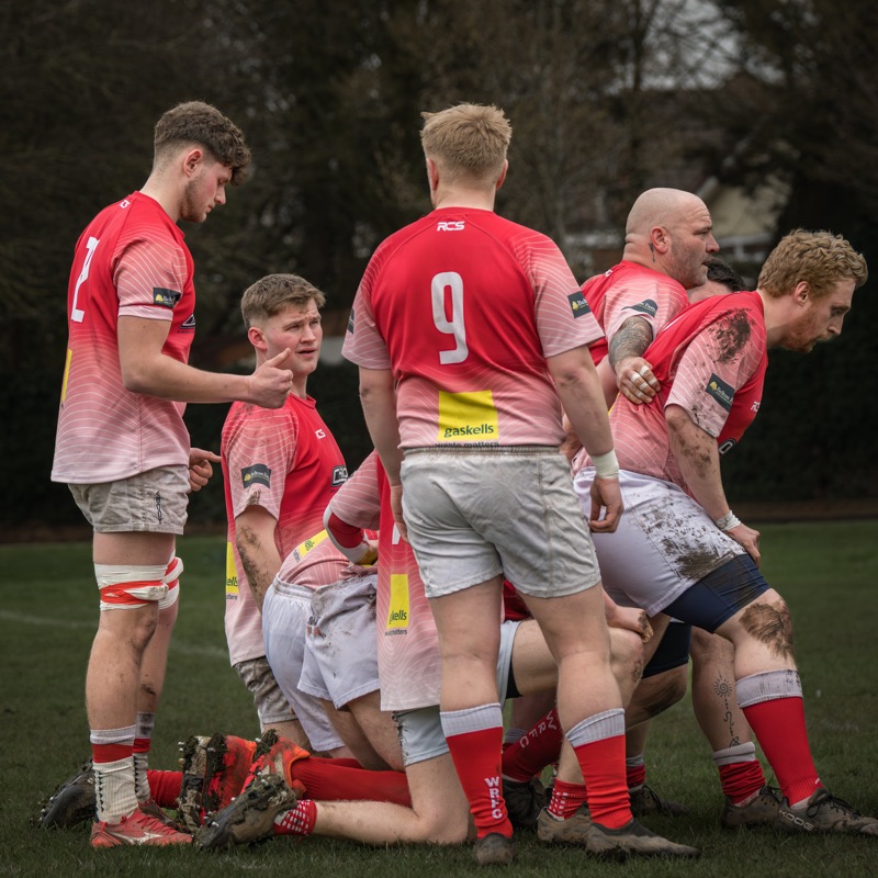A group of rugby players, wearing red jerseys and white shorts, are huddled together on a rugby field during daytime. They are kneeling with their heads bowed, possibly during a team prayer or moment of reflection.