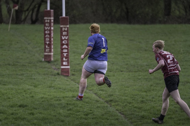 Two rugby players on a grassy field near the try line, one running towards the goalpost and the other in pursuit. The player in blue has red hair and is wearing a blue jersey with the number 1, while the other player has light brown hair and is weari