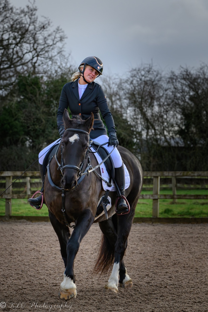 A woman riding a black and white horse in an outdoor riding arena on a cloudy day, with trees and a wooden fence in the background.