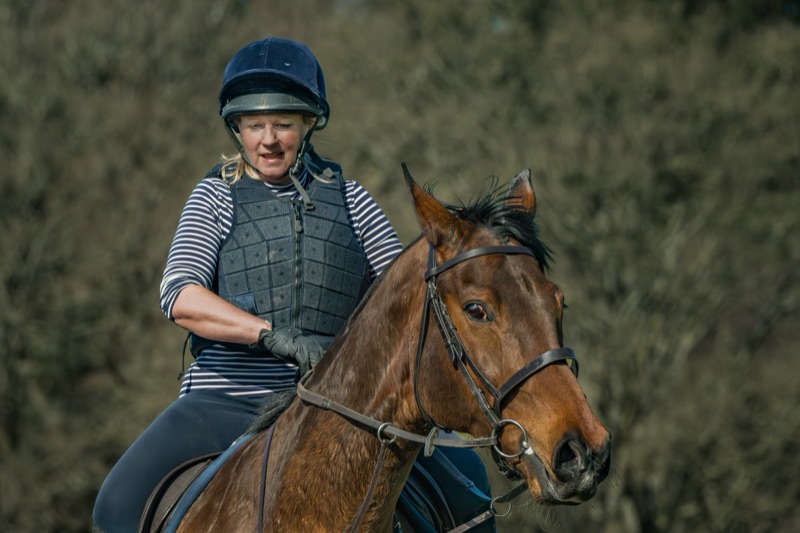 A woman wearing a helmet, striped shirt, and protective vest riding a brown horse with a black mane outdoors with blurred greenery background.