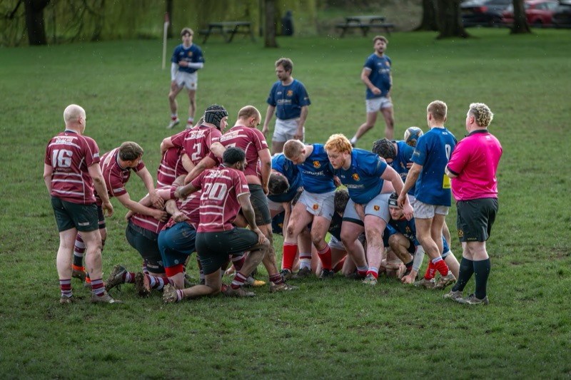 Rugby players engaged in a scrum during a match on a grassy field, with some players wearing team jerseys and one referee observing.