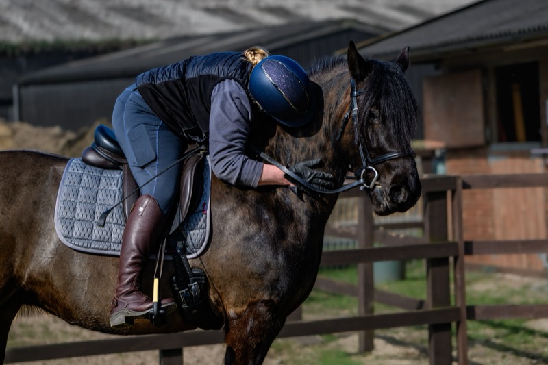Person wearing riding gear, helmet, and gloves leaning forward on a dark brown horse with a black mane, in a riding arena with a wooden fence and a building in the background.