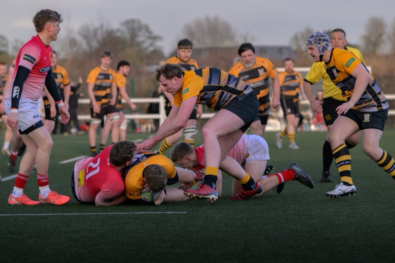 A rugby match with players from two teams, one in yellow and black uniforms and the other in red and white uniforms, engaged in a tackle on the field during daylight. Some players are on the ground and others are standing around, watching and partici