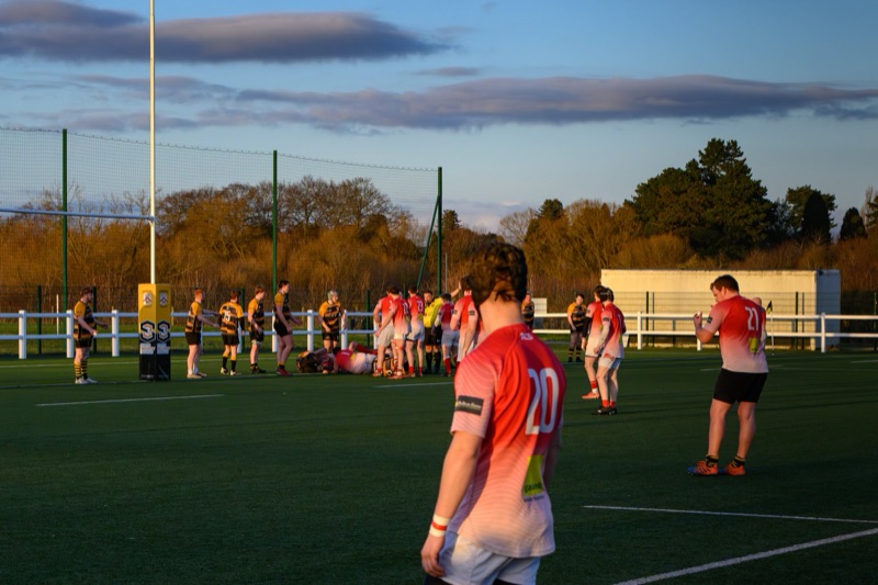 A rugby match taking place on an outdoor field during sunset, with players gathered around the ground and one player lying on the grass, with a backdrop of trees and a partly cloudy sky.