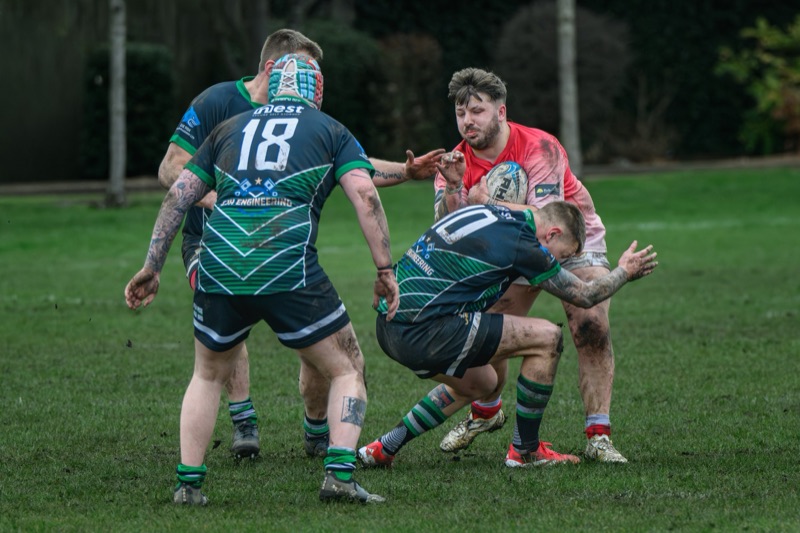 Rugby players engaged in a tackle during a match on a grassy field with trees in the background.