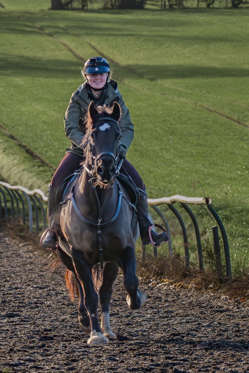 A person riding a horse on a dirt path in a green field, wearing a helmet and jacket, during daytime.