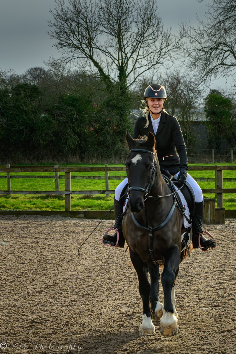 A smiling woman riding a horse in an outdoor riding arena, wearing a black riding helmet, black jacket, and white riding pants. The horse is dark-colored with white markings on its legs, and the background shows bare trees and a wooden fence.