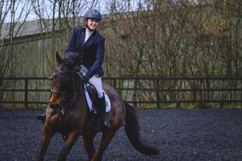 A young woman riding a horse in an outdoor riding arena, wearing a helmet and riding attire, with trees and a fence in the background.