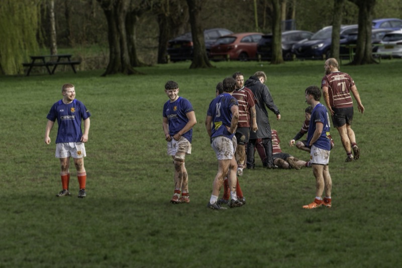 A group of rugby players standing and sitting on a muddy field after a match, with some players smiling and others looking serious, surrounded by trees and parked cars.