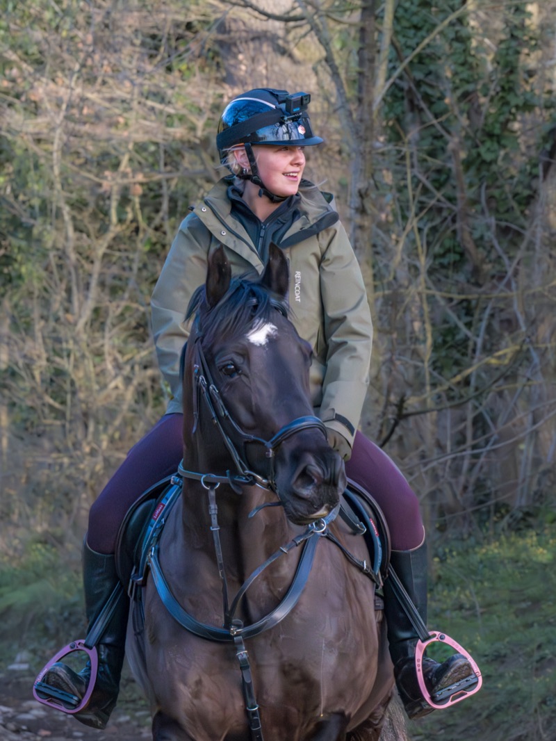 A woman riding a horse outdoors, dressed in a brown jacket and wearing a helmet with a camera mounted on top, surrounded by trees and foliage.
