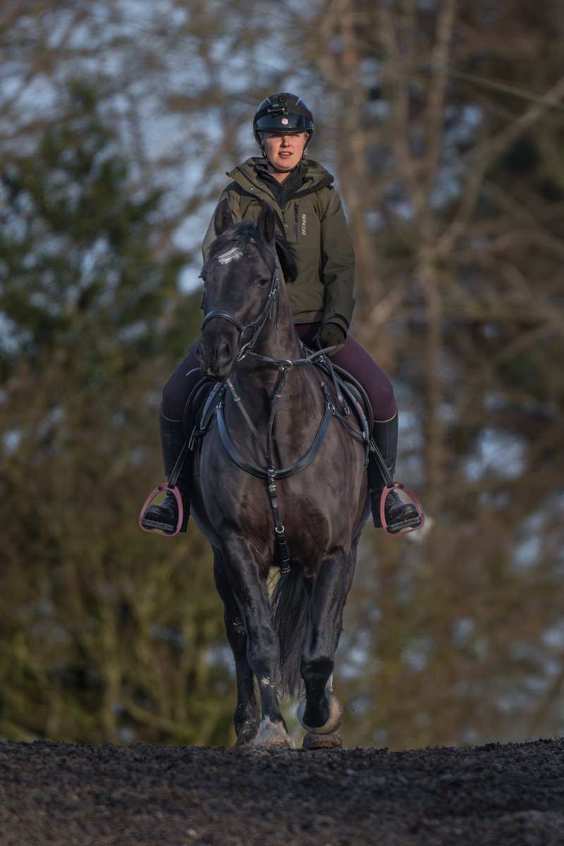 A woman riding a gray horse outdoors, wearing a black helmet with a camera mounted on top, a green jacket, and dark riding pants. The background shows blurred trees.