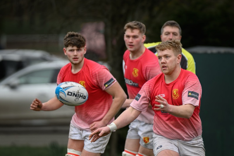 Four rugby players in red jerseys running on the field, with two of them holding a rugby ball, during a match.