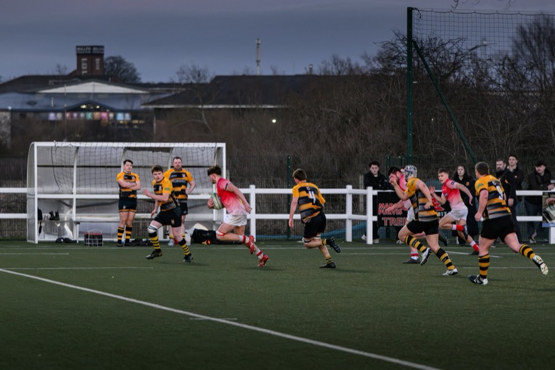 Rugby players on a field during a game, with some running and others standing near the sideline, with trees, buildings, and a cloudy sky in the background.