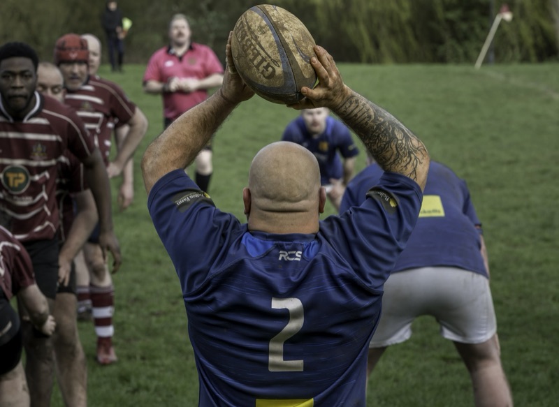 A rugby player in a blue jersey prepares to throw a rugby ball during a game, with team members and a coach watching on the sideline.