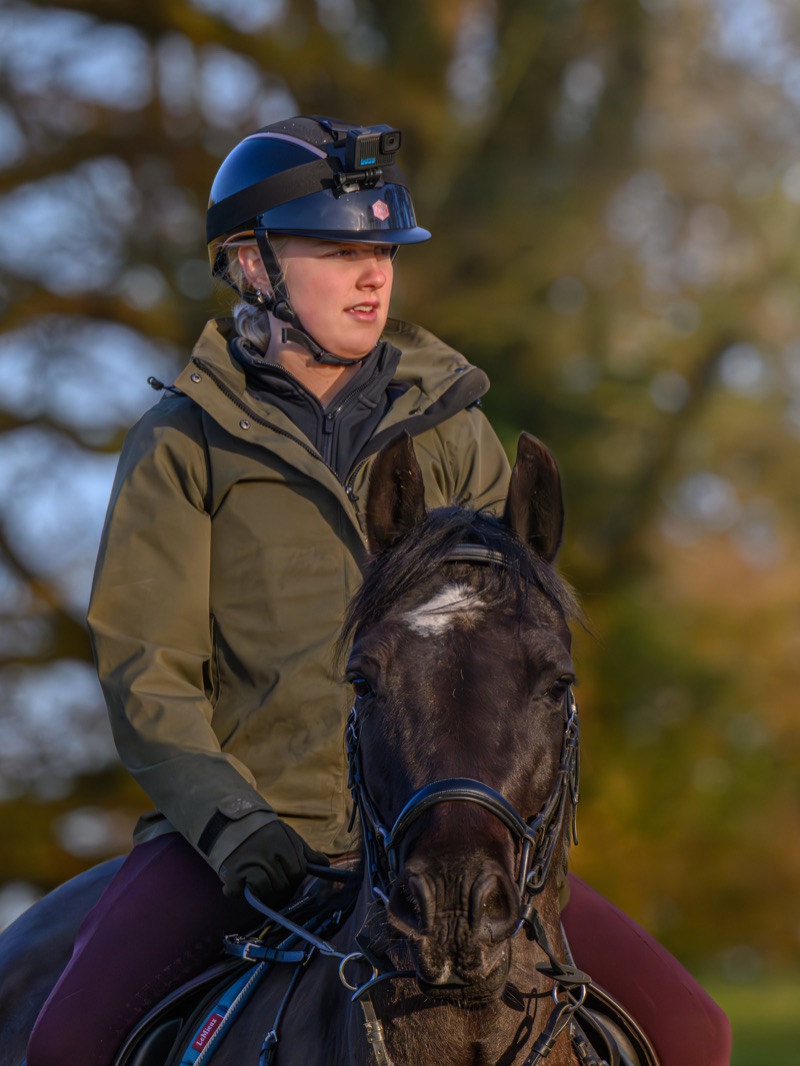 A woman wearing a helmet with a camera mounted on it, riding a horse outdoors during autumn.