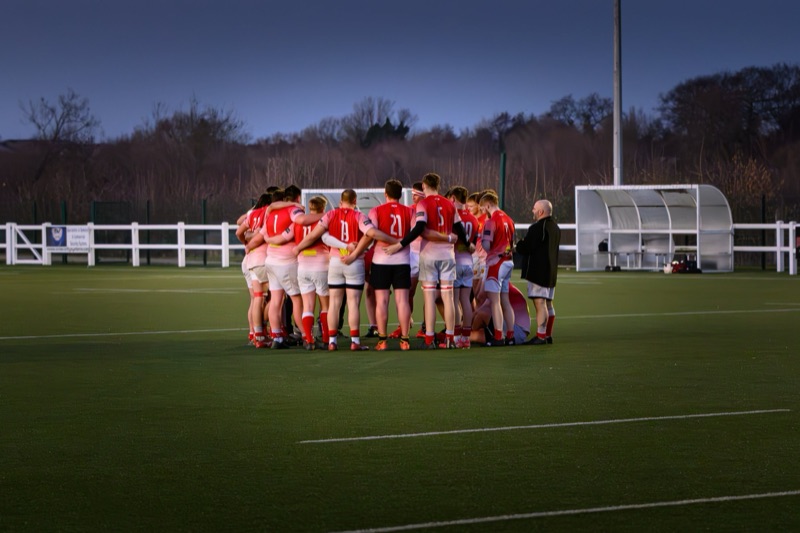 A group of rugby players huddle together on a rugby field during twilight, with a coach standing nearby. They are wearing red uniforms and are standing in a circle with arms around each other's shoulders.