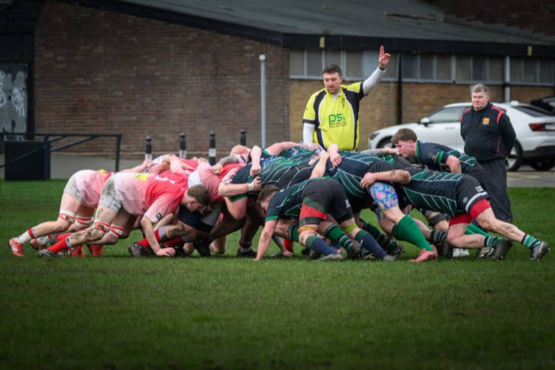 A group of rugby players in a scrum on a grassy field, with a coach or referee overseeing. Two men are standing behind the scrum, one raising his hand.