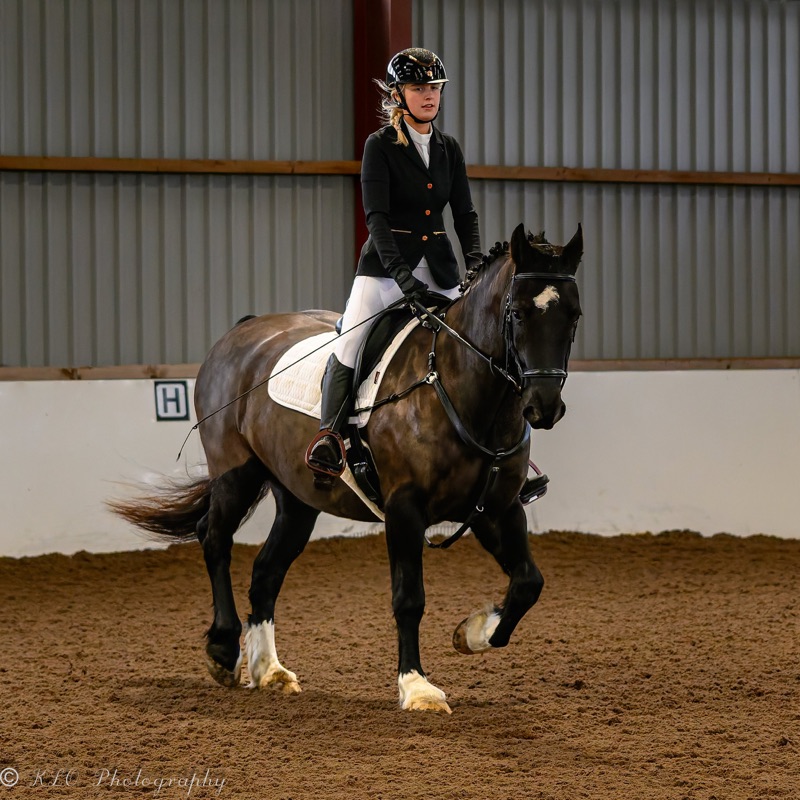 A young woman riding a brown and white horse inside an indoor riding arena with a corrugated metal wall in the background.