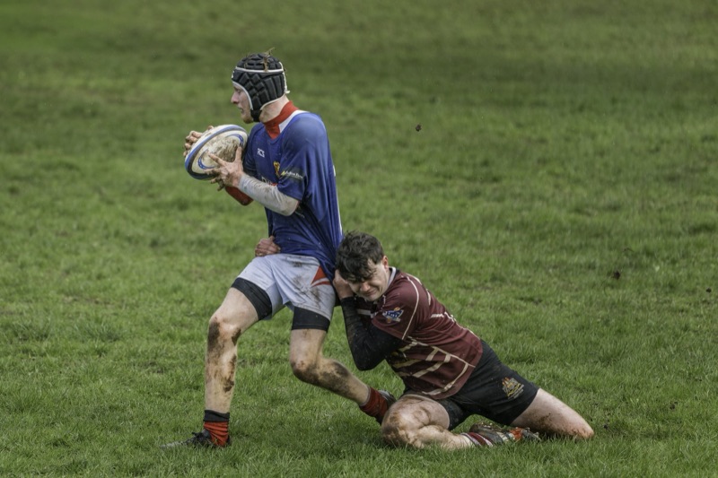 Two rugby players engaging in a tackle on a muddy grass field during a game, with grass and mud on their bodies.