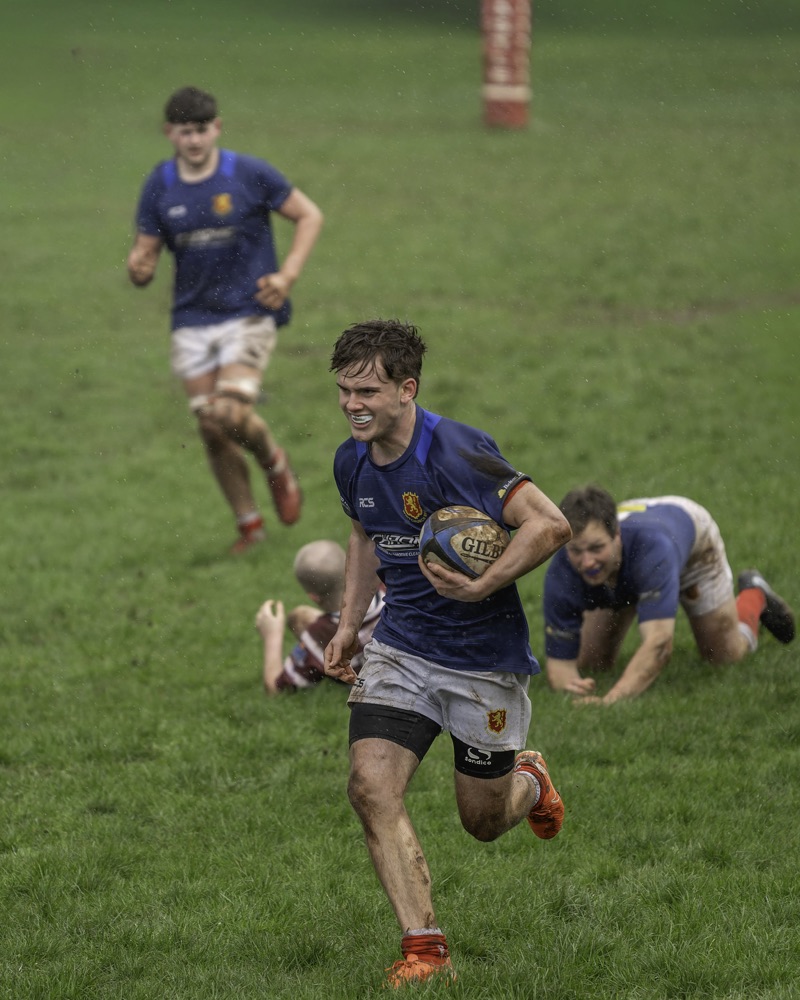 Rugby players on a muddy field during a match, with one player running with the ball in the foreground, and others in the background chasing or on the ground.