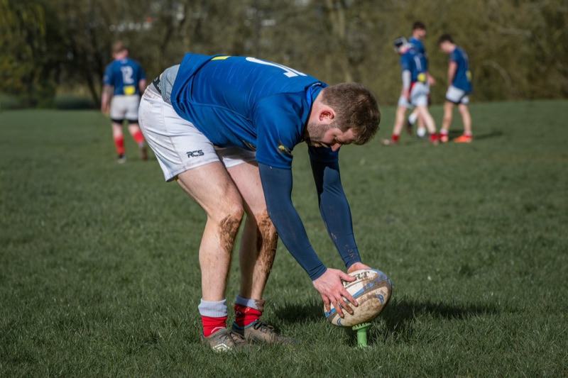 A man playing rugby on a grassy field, bent over and placing a rugby ball on a kicking tee, with four players in blue jerseys standing in the background.