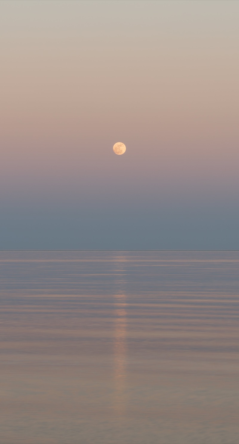 Moon rising over a calm ocean during dusk with pastel-colored sky.