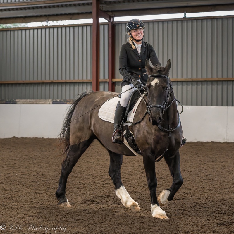 A woman riding a horse inside an indoor riding arena with a smiling expression, wearing a black helmet, black riding jacket, and white riding pants.
