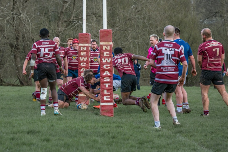 Rugby players in maroon jerseys on the field, competing near the goal post as some players reach for the ball.