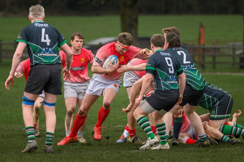 Rugby players in a scrum during a game on a grassy field, with players wearing red and black jerseys with green-striped socks, and black jerseys with blue accents, some muddy, with trees in the background.
