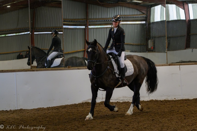 A woman riding a horse inside an indoor equestrian arena, with her reflection visible in large mirrors on the wall.