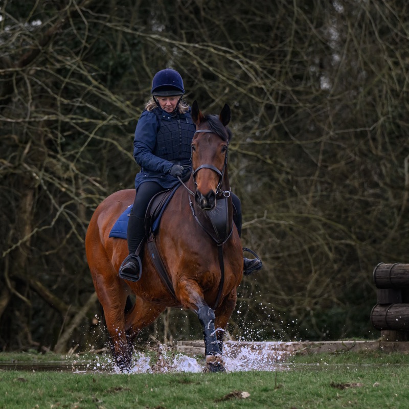 A woman wearing riding gear, including a helmet, riding a brown horse through a water obstacle outdoors with trees in the background.