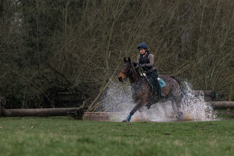A woman riding a horse through water, wearing a helmet and riding gear, with dense trees in the background.