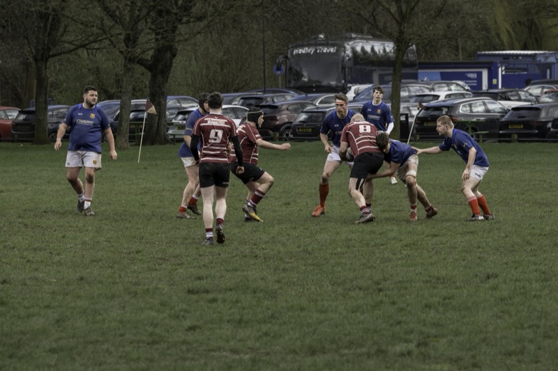 Group of rugby players in a match, some tackling and others observing, on a grassy field with parked cars and trees in the background.