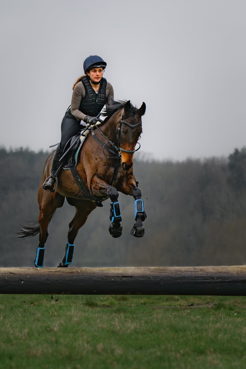 A woman wearing a helmet, a protective vest, and gloves riding a galloping horse over a wooden obstacle during a competition or training session outdoors.