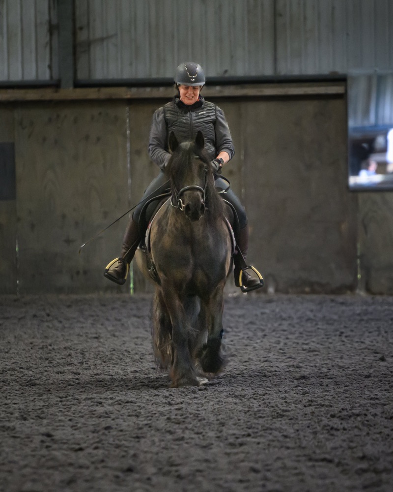 A person riding a horse in an indoor riding arena, wearing a black helmet, black gloves, a gray jacket, and black riding pants. The horse is dark-colored and appears to be trotting towards the camera. The background shows wooden walls and a small win