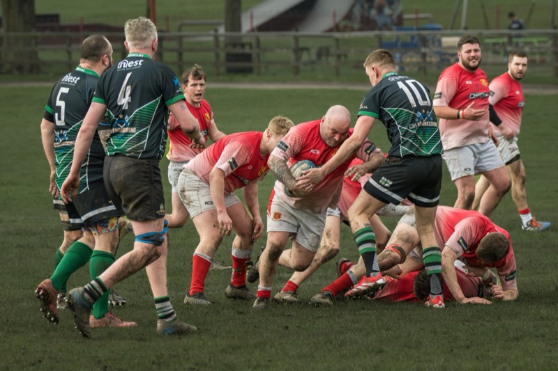 Rugby players in red and black uniforms compete on the field with one player holding the ball, while others push and tackle during a game in an outdoor stadium.