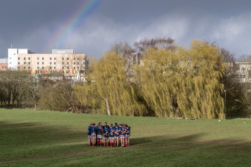 A group of young soccer players in blue uniforms and a coach standing in a circle on a grassy field, with trees and a building with a rainbow in the sky in the background.