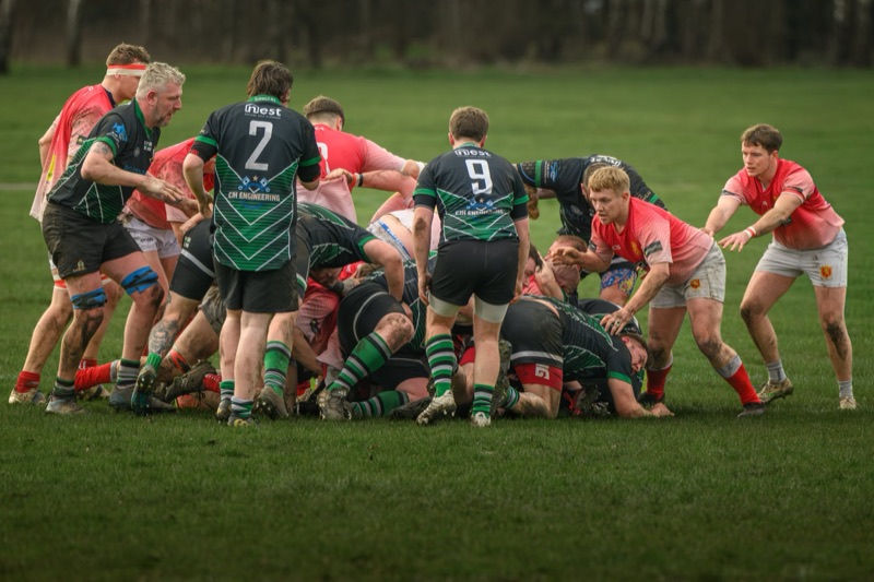 Rugby match in progress with players engaged in a ruck on a grassy field, some are wearing black jerseys with green accents, others are in red and pink jerseys.