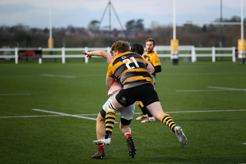 Two rugby players competing for the ball on a green field, with a border fence and cloudy sky in the background.